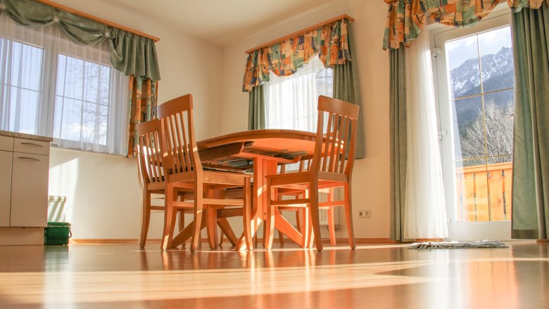 Apartment 70m², © Haus Lenz Dining area with wooden table and chairs in a sunny room with large windows and curtains.