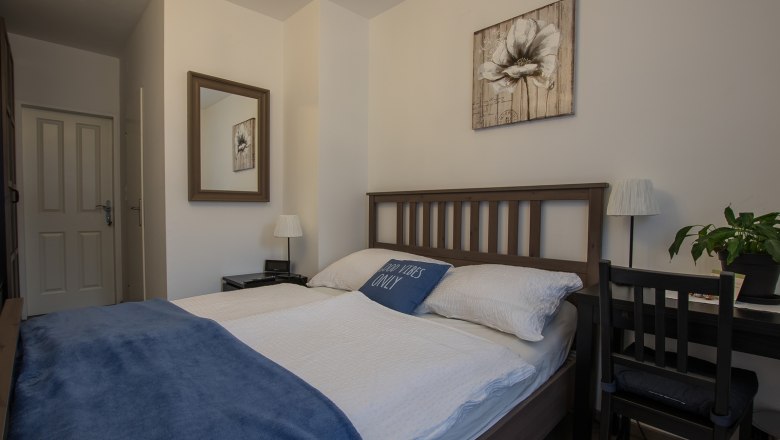 Doppelzimmer Fuchs, © Dirnbauer Gemütliches Doppelzimmer mit Holzbett, blauer Decke und Dekokissen. Ein Spiegel und ein Blumenbild an der Wand, daneben ein Schreibtisch mit Pflanze.