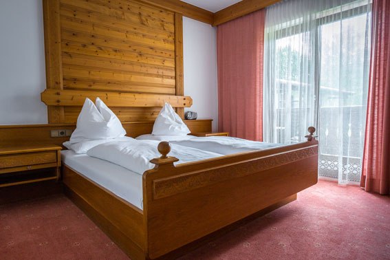 Sleep comfortably at the Bruckerhof, © Wiener Alpen / Christian Kremsl A cozy bedroom with a wooden double bed, white comforters and red curtains in front of a window.