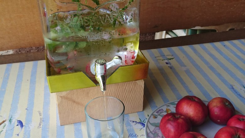 Refreshing spring water, © Scheibenreif Karin A glass container with water and herbs, next to it a glass and a plate with red apples on a striped table.
