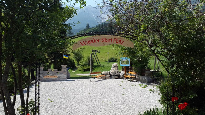 Hiking starting point, © Forellenhof Entrance to a hiking starting point with benches and a sign, surrounded by trees and mountains in the background.