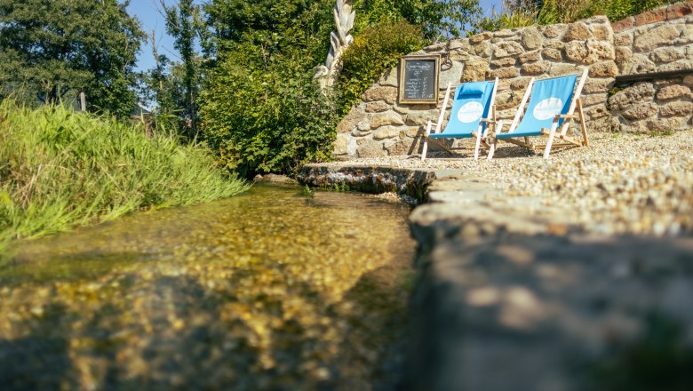 Sommerfrische beim Gasthof Schmirl, © Martin Fülöp Zwei blaue Liegestühle stehen neben einem kleinen Bach vor einer Steinmauer.