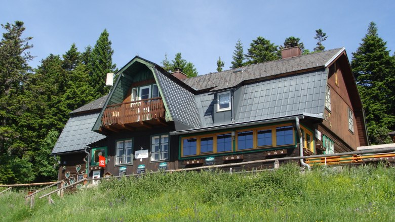 Hubertus House, © OEAV Gebirgsverein, Foto Robert Vondracek A traditional mountain house with a wooden façade and shingle roof, surrounded by trees and meadows.