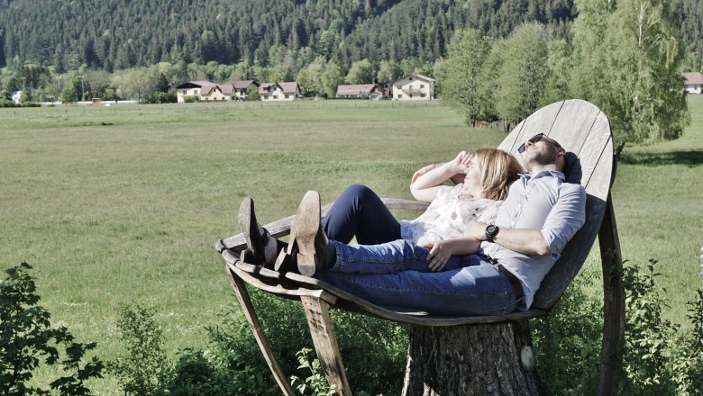 Kuschelliege, © Gasthof Schmirl Ein Paar liegt auf einer großen Holzliege in einer grünen Landschaft mit Bergen im Hintergrund.