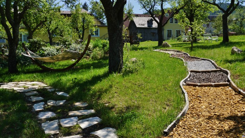 Barfussweg, © Gasthof Schmirl Ein Barfußweg mit verschiedenen Materialien in einem grünen Garten mit Bäumen und einer Hängematte.