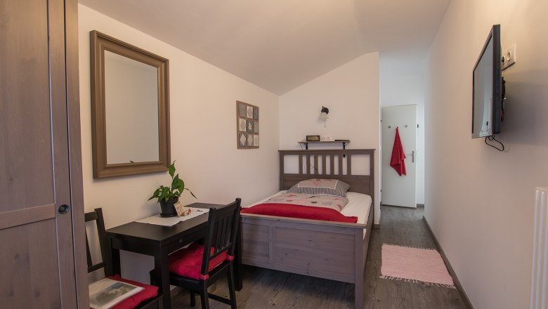 Doppelzimmer Wolf, © A. Dirnbauer Ein gemütliches Einzelzimmer mit Holzmöbeln, einem Bett, einem kleinen Tisch mit Stühlen, einem Spiegel und einem Fernseher an der Wand.