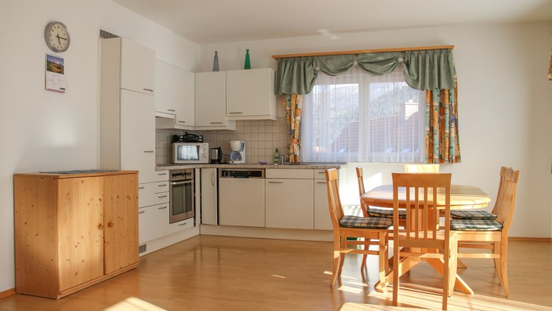 Apartment 70m², © Haus Lenz Bright kitchen with dining table and chairs, white cupboards, wooden floor, windows with curtains.