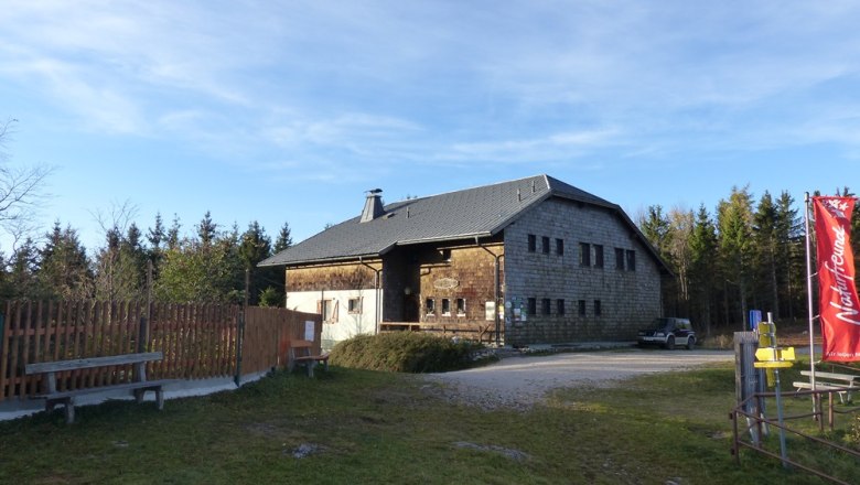Öhler Schutzhaus, © Mario Putz Ein rustikales Gebäude mit Holzwänden und einem grauen Dach, umgeben von Bäumen. Eine rote Fahne steht rechts im Bild.