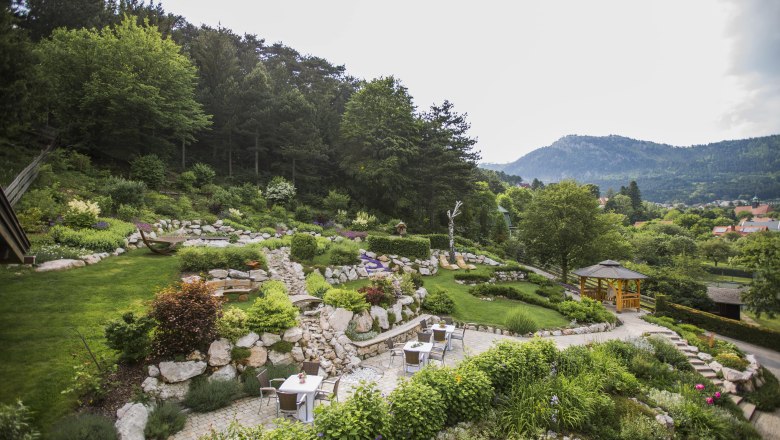 Ein terrassierter Garten mit Steinen, Pflanzen und Sitzbereichen, umgeben von B&auml;umen und Bergen im Hintergrund.