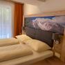 A cozy double room with a large bed, wooden furniture and a picture of mountains above the headboard. A window with curtains can be seen.