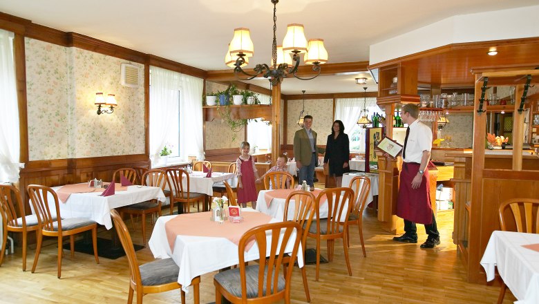 Ein rustikales Restaurant mit Holzm&ouml;beln und Tischdecken. Ein Kellner begr&uuml;&szlig;t eine Familie am Eingang.