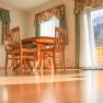 Dining area with wooden table and chairs in a sunny room with large windows and curtains.