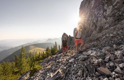 Zwei Wanderer mit Rucksäcken steigen auf steinigem Gebirgspfad bei Sonnenschein und Bergpanorama auf