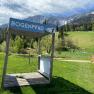 Holzplattform mit Schild 'Bogenpfad' vor einer Berglandschaft.
