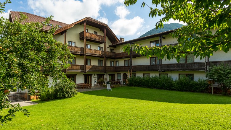 Ein traditionelles Hotel mit Holzbalkonen und einem gepflegten Garten im Vordergrund, umgeben von B&auml;umen und Bergen im Hintergrund.