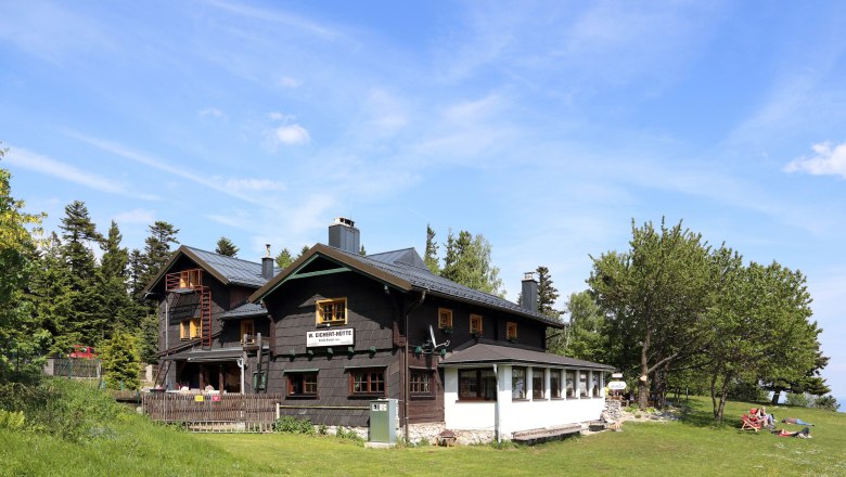 Wilhelm Eicherthütte, © BwagCommons Eine Berghütte mit dunklem Holz und weißen Fenstern, umgeben von Bäumen und Wiese, unter blauem Himmel.