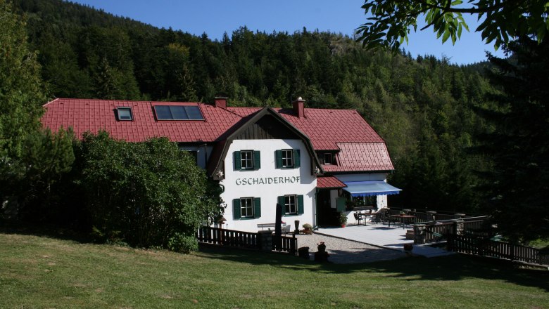 Ein traditionelles Haus mit rotem Dach, umgeben von Wald und Wiese, mit der Aufschrift 'Gschaiderhof'.