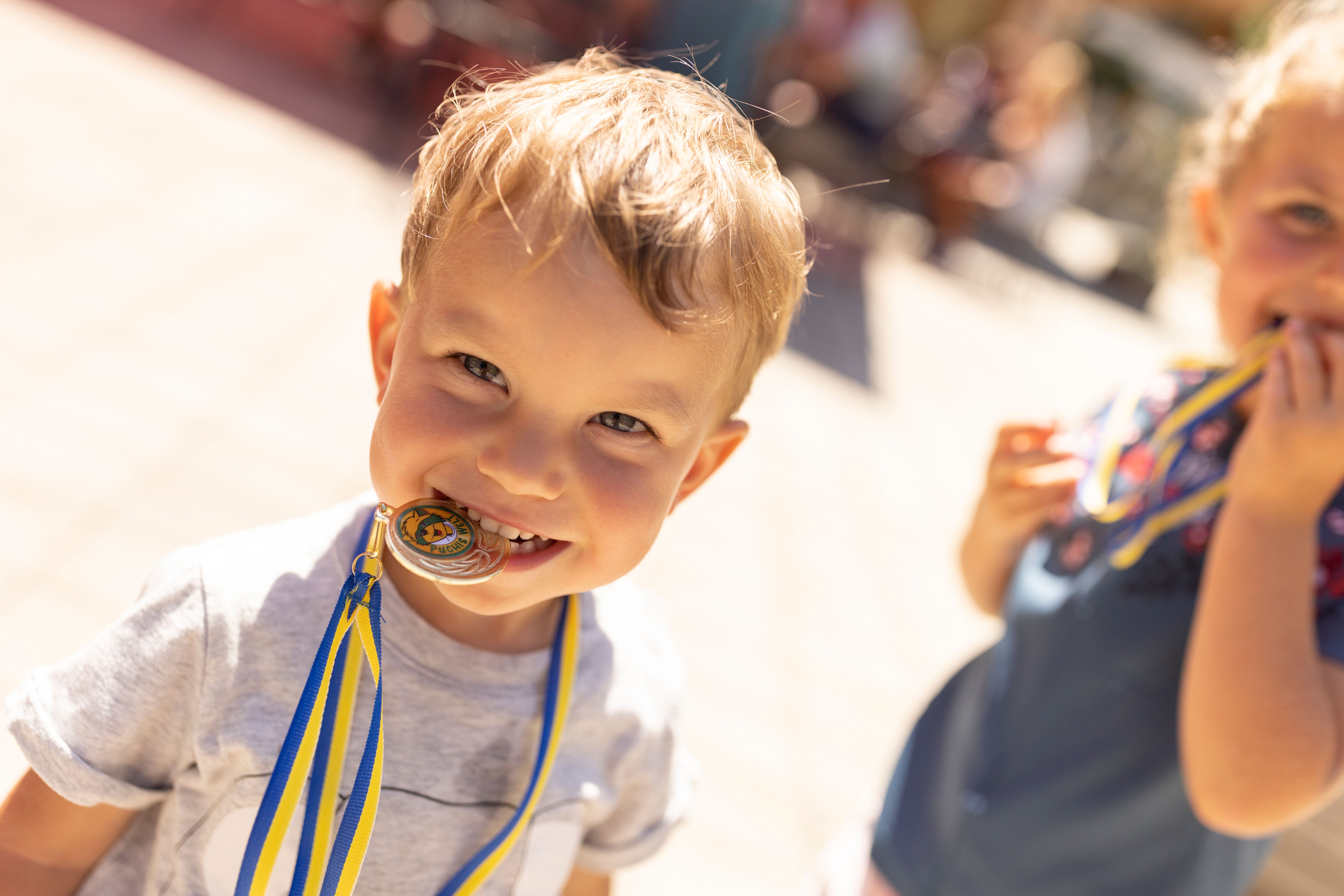 Junge bei Sonnenschein beißt lachend auf eine Medaille mit blau-gelbem Band, im Hintergrund ein weiteres Kind unscharf sichtbar