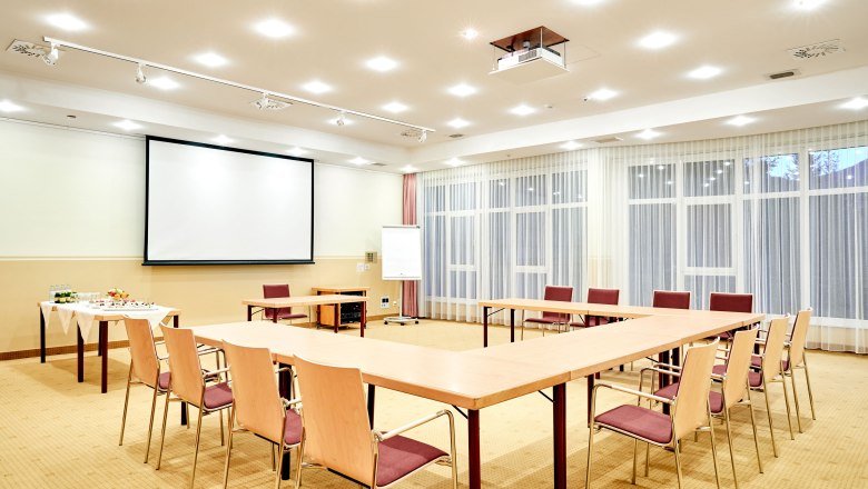 Ein heller Seminarraum mit U-f&ouml;rmiger Tischanordnung, Leinwand, Flipchart und Getr&auml;nketisch.