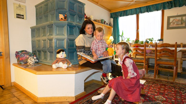 Eine Frau sitzt mit zwei Kindern auf einer Bank vor einem Kachelofen und liest ein Buch. Spielzeug und Dekorationen sind um sie herum verteilt.