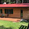 Ein kleines Holzferienhaus mit rotem Dach, Terrasse und Gartenm&ouml;beln, umgeben von Wald.