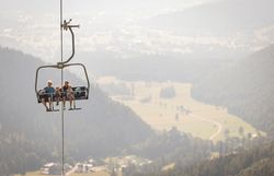 Vier Personen sitzen auf einem Sessellift über bewaldeten Hügeln mit weiter Landschaft im Hintergrund
