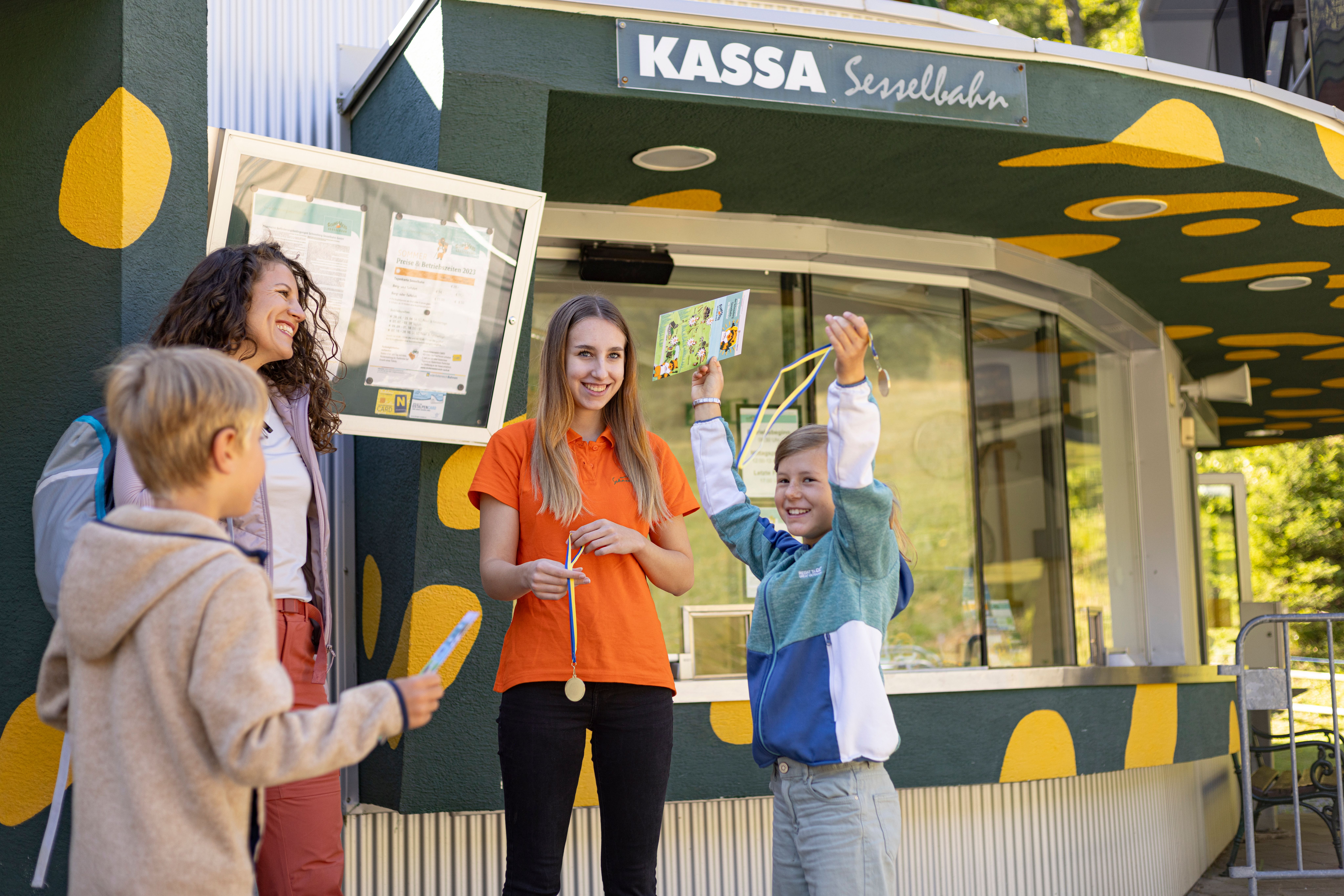Vier Personen vor einer Kasse mit der Aufschrift 'KASSA Sesselbahn', zwei Kinder halten bunte Tickets, eine Frau und ein Mädchen lächeln.