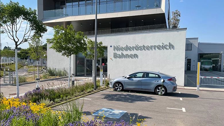 Gebäude mit der Aufschrift »Niederösterreich Bahnen«, davor ein geparkter silberner Pkw und bepflanzte Flächen mit Blumen und Büschen