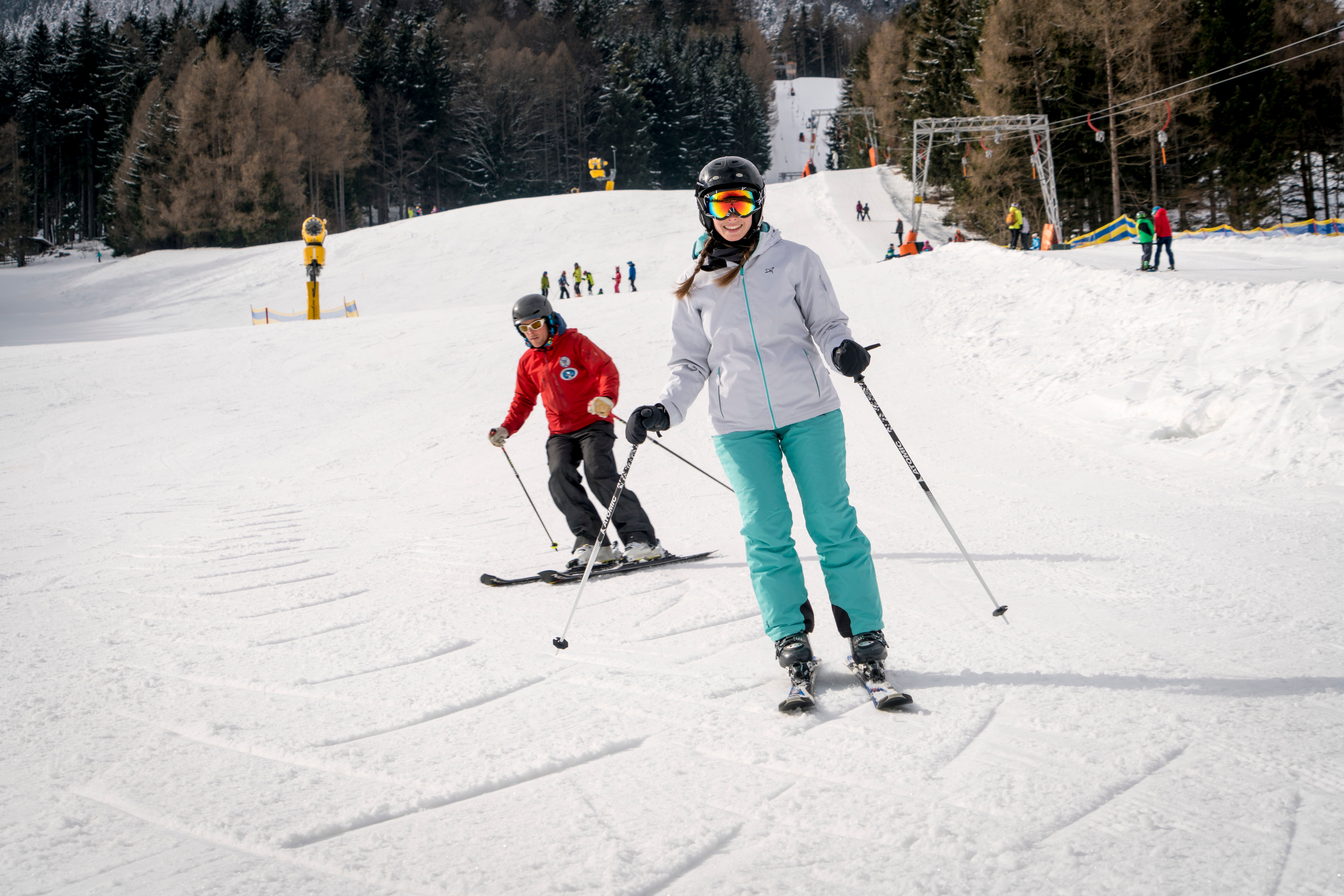 Zwei Skifahrer in Winterkleidung fahren auf einer präparierten Skipiste mit Wald im Hintergrund