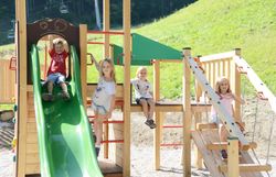 Vier Kinder spielen auf einem Holzspielplatz mit grüner Rutsche, Klettergerüst und Schaukel im Freien auf einer Wiese.