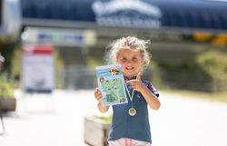 Mädchen mit Medaille um den Hals hält eine Karte und zeigt darauf, im unscharfen Hintergrund die blaue Talstation mit weißer Aufschrift