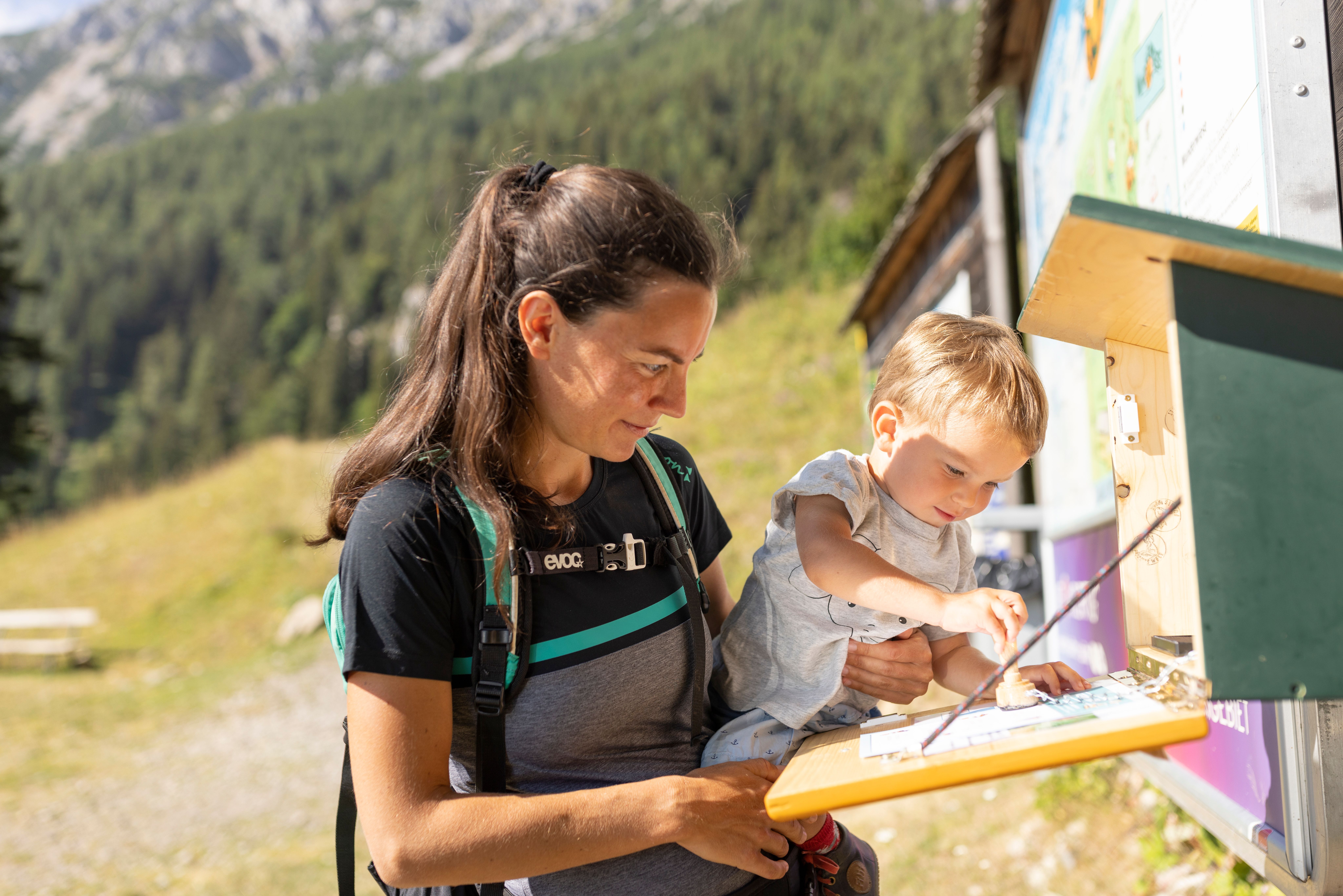 Frau mit Pferdeschwanz und Rucksack hält Kleinkind, das an einer Infotafel im Gebirge zeigt