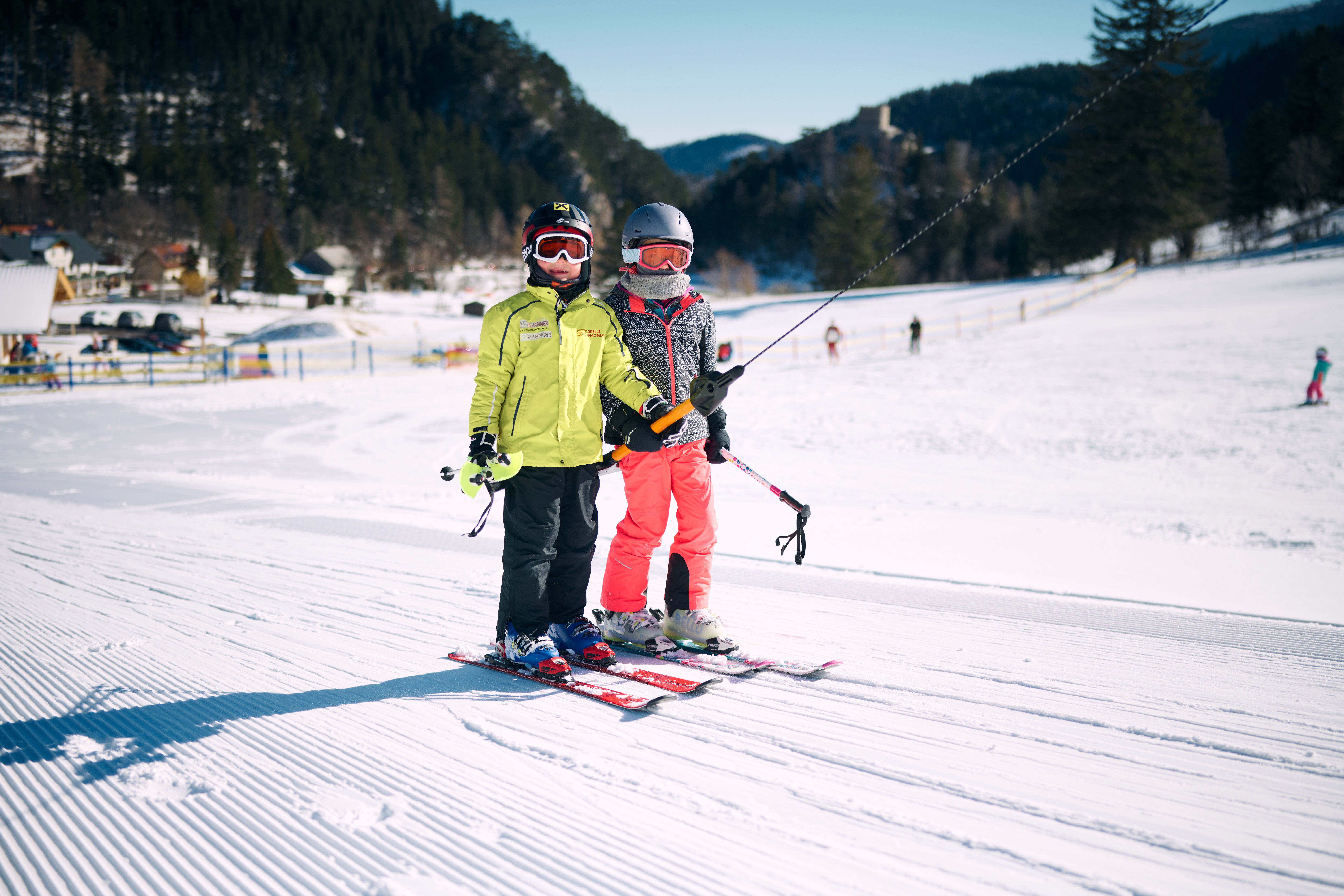 Zwei Kinder in Skibekleidung mit Helm und Skibrille stehen nebeneinander auf Skiern auf einer präparierten Skipiste im Winter