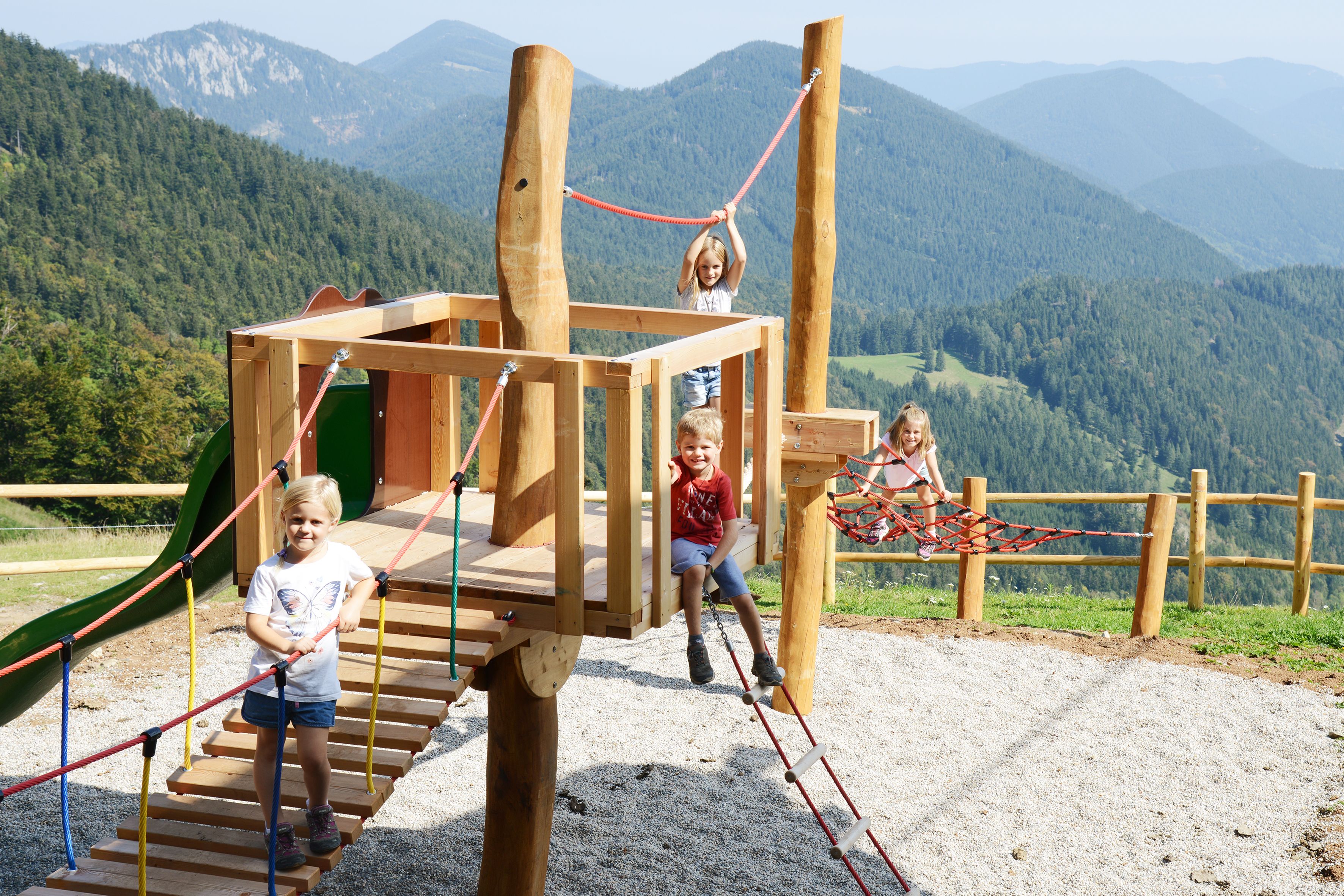 Vier Kinder spielen auf einem Holz-Klettergerüst mit Seilbrücke vor bewaldeten Bergen und blauem Himmel
