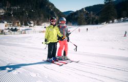 Zwei Kinder in Skibekleidung mit Helm und Skibrille stehen nebeneinander auf Skiern auf einer präparierten Skipiste im Winter