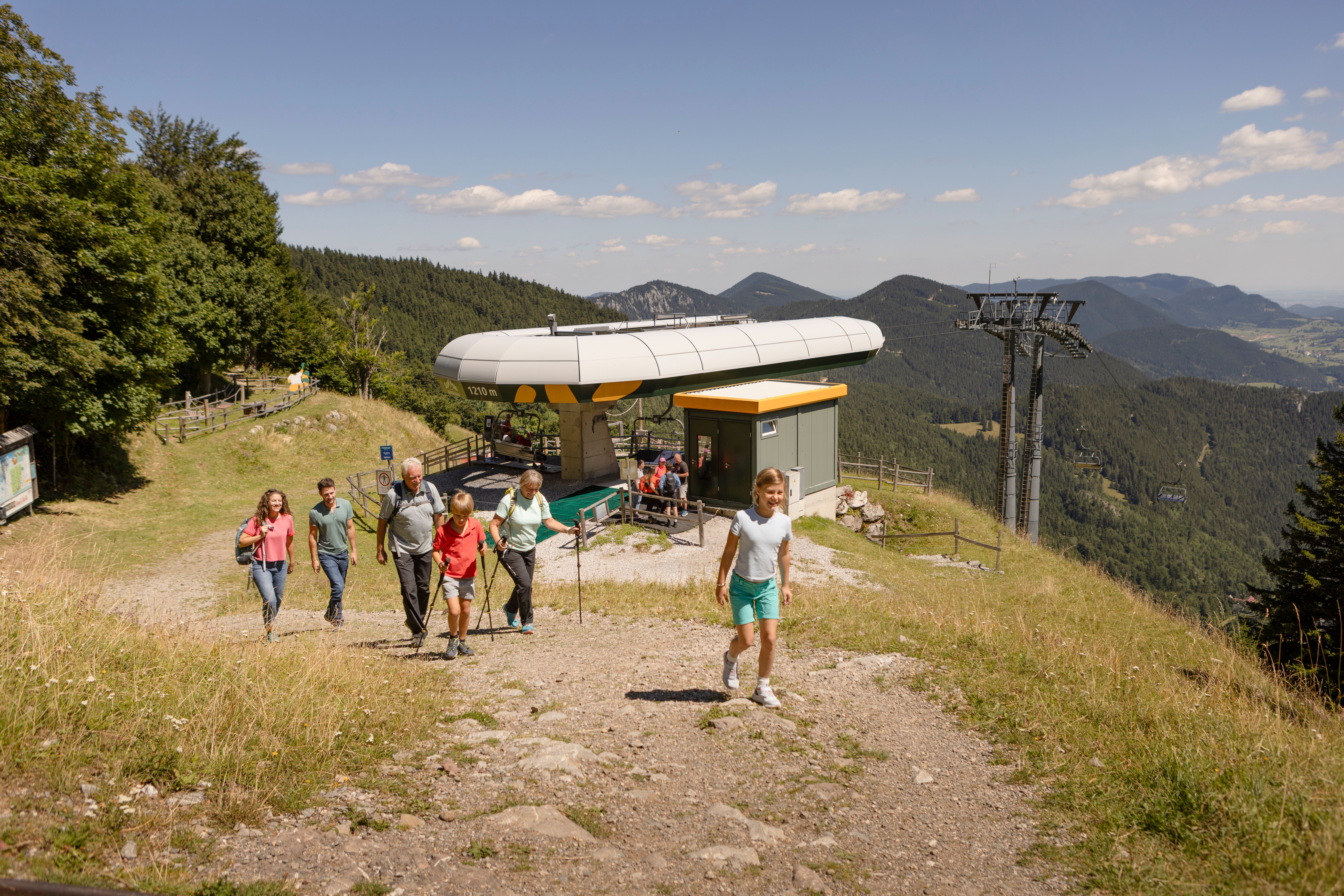 Eine Gruppe wandert auf einem steinigen Bergweg an einer Seilbahnstation vorbei. Im Hintergrund sind bewaldete Berge und ein Tal zu sehen.