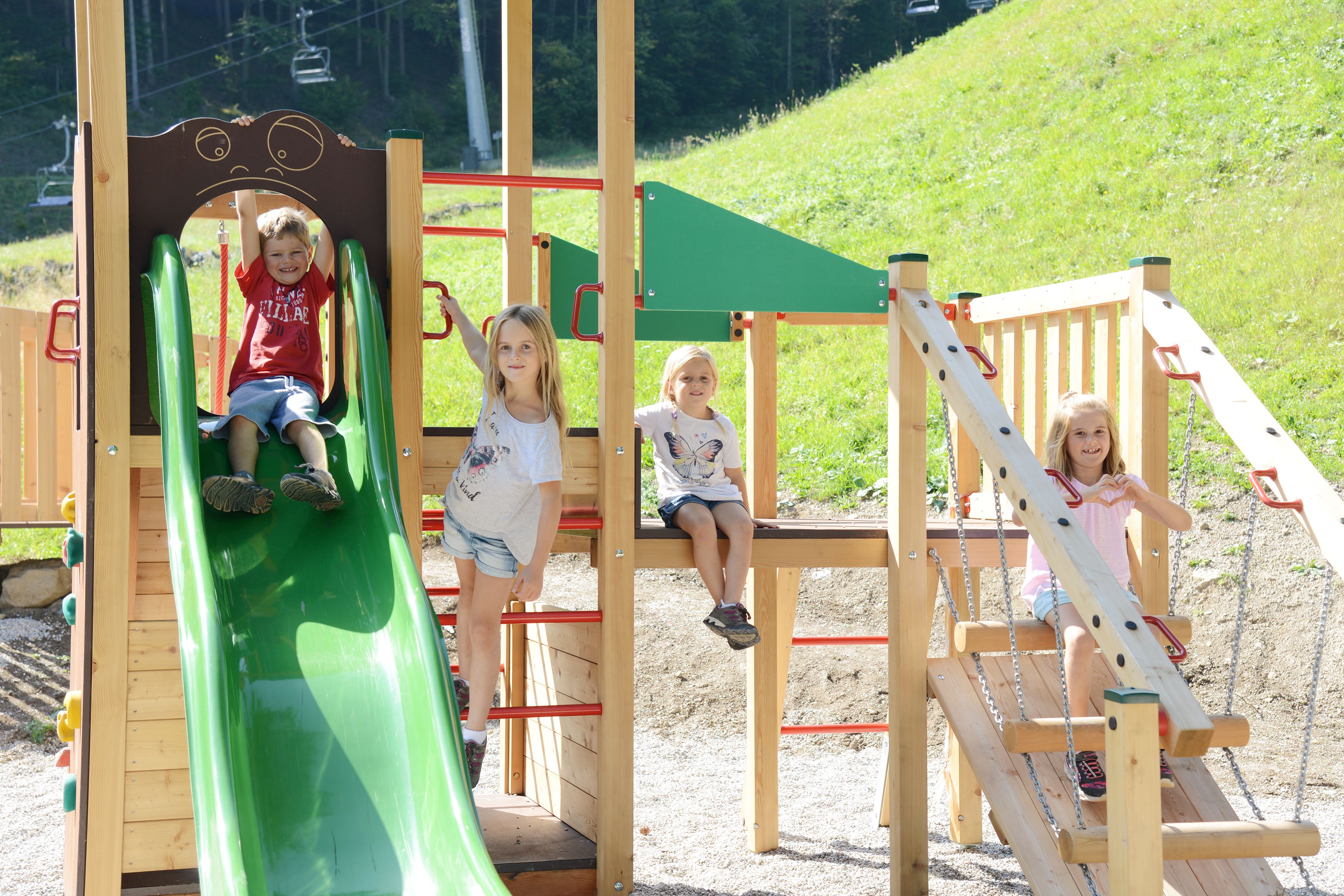Vier Kinder spielen auf einem Holzspielplatz mit grüner Rutsche, Klettergerüst und Schaukel im Freien auf einer Wiese.