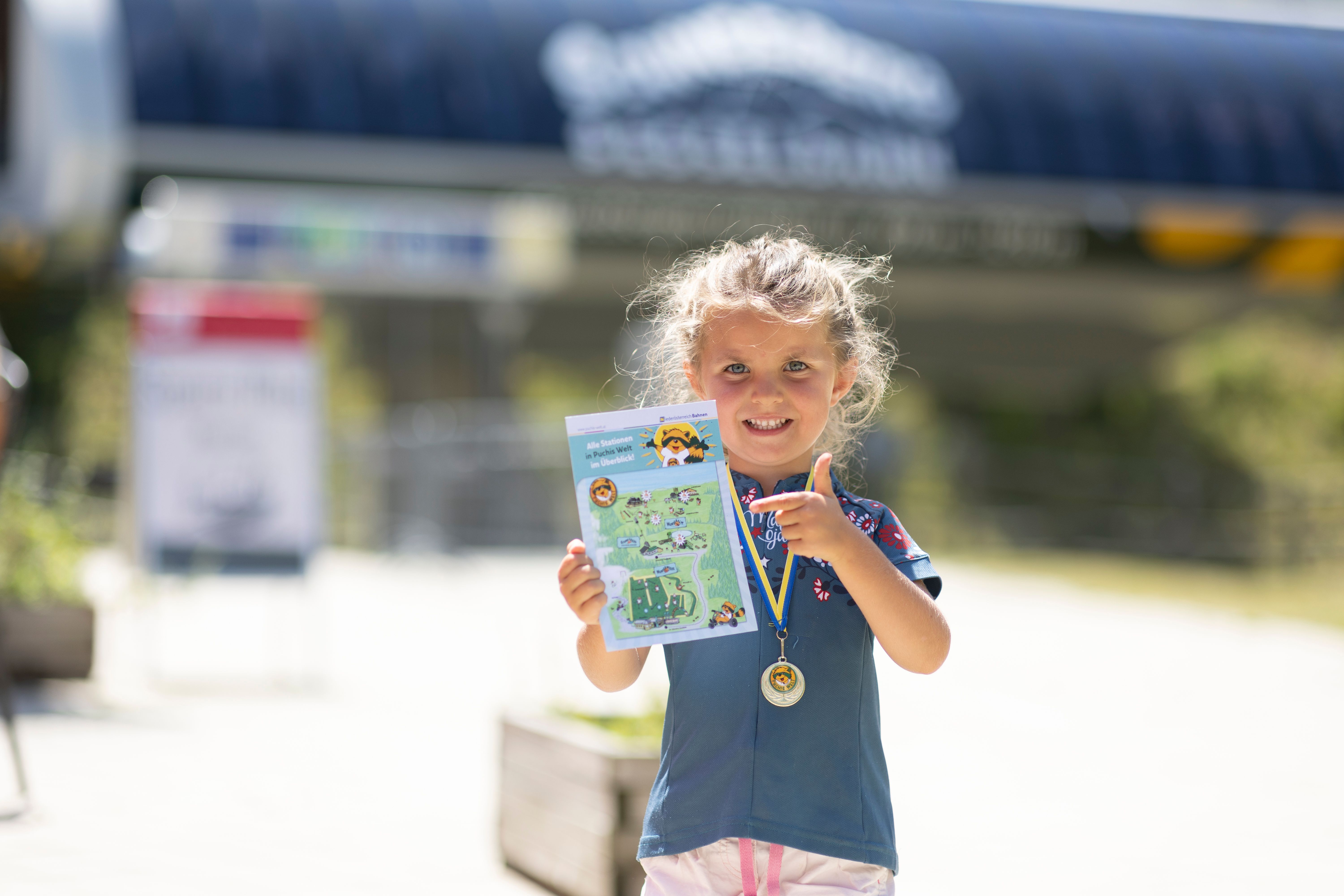 Mädchen mit Medaille um den Hals hält eine Karte und zeigt darauf, im unscharfen Hintergrund die blaue Talstation mit weißer Aufschrift