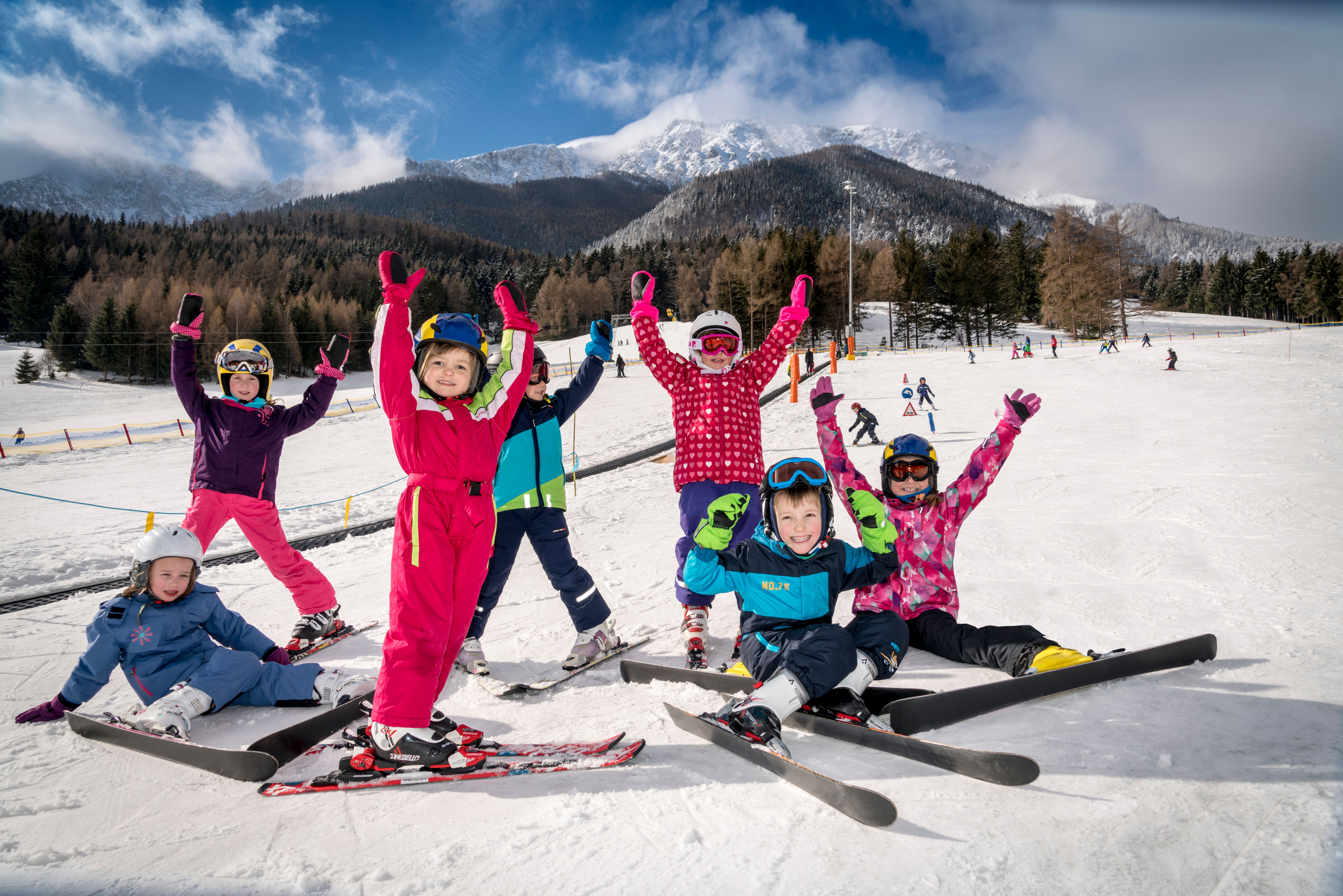 Sieben Kinder in bunter Skibekleidung und Helmen posieren auf einer schneebedeckten Piste vor bewaldeten Bergen und blauem Himmel, einige mit erhobenen Armen