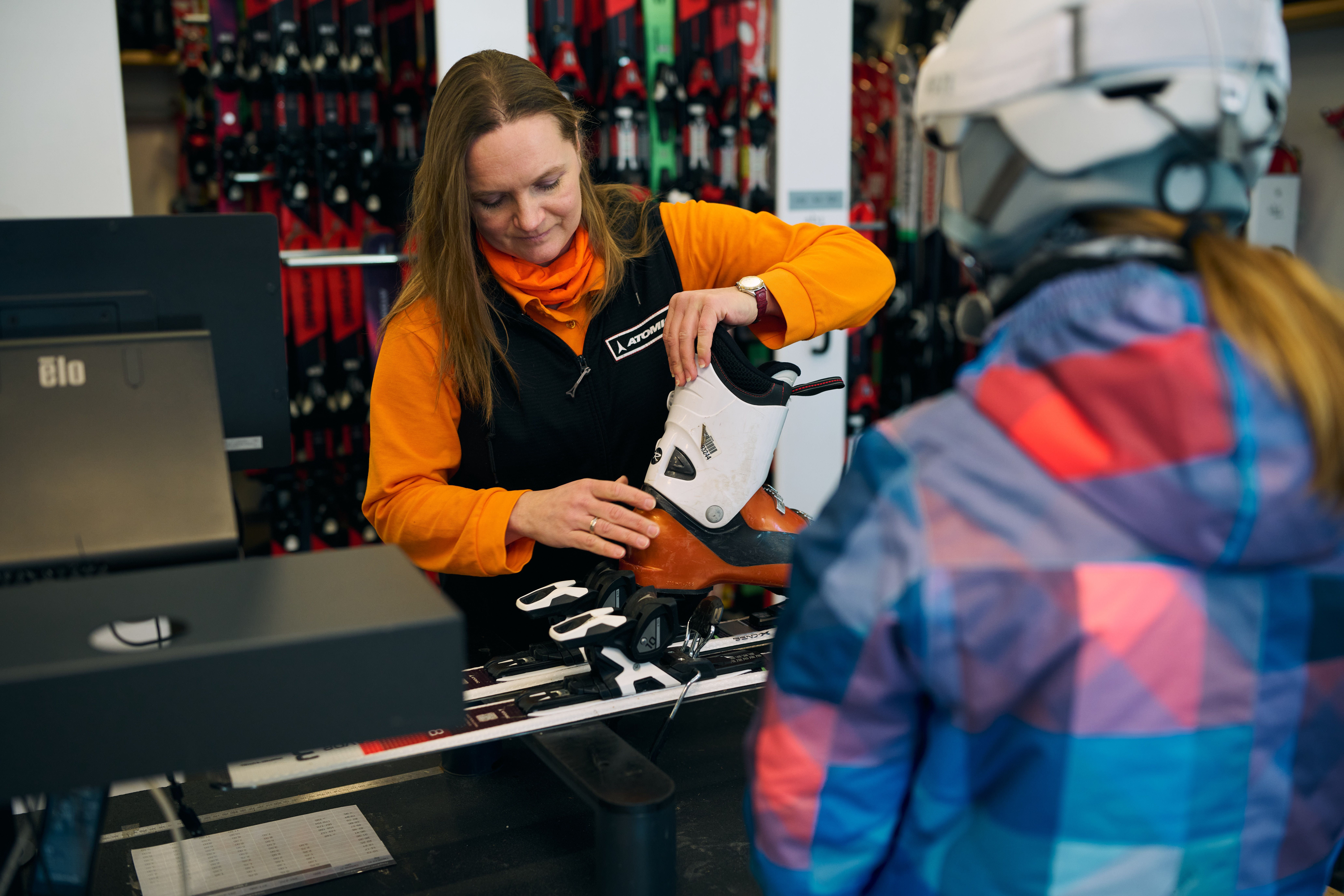 Verkäuferin mit orangefarbenem Pullover montiert Skischuh an Skibindung vor Kundin in Skihelm und bunter Jacke