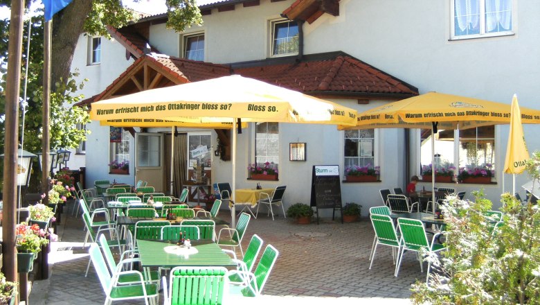 Ein Gastgarten mit gr&uuml;nen St&uuml;hlen und gelben Sonnenschirmen vor einem Geb&auml;ude.