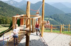 Vier Kinder spielen auf einem Holz-Klettergerüst mit Seilbrücke vor bewaldeten Bergen und blauem Himmel