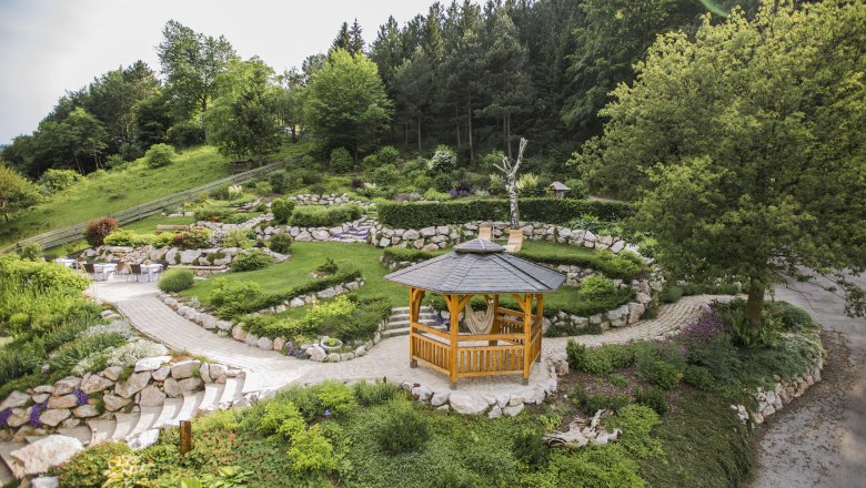 Ein h&ouml;lzerner Pavillon in einem gepflegten Garten mit gr&uuml;nen B&auml;umen und Steinwegen.