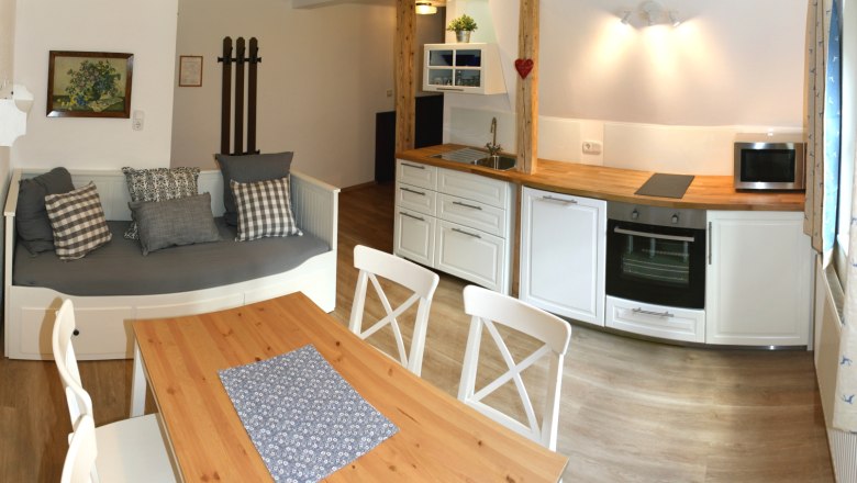 Gem&uuml;tliche K&uuml;che mit Holztisch, wei&szlig;en St&uuml;hlen, Sofa und moderner K&uuml;chenzeile.