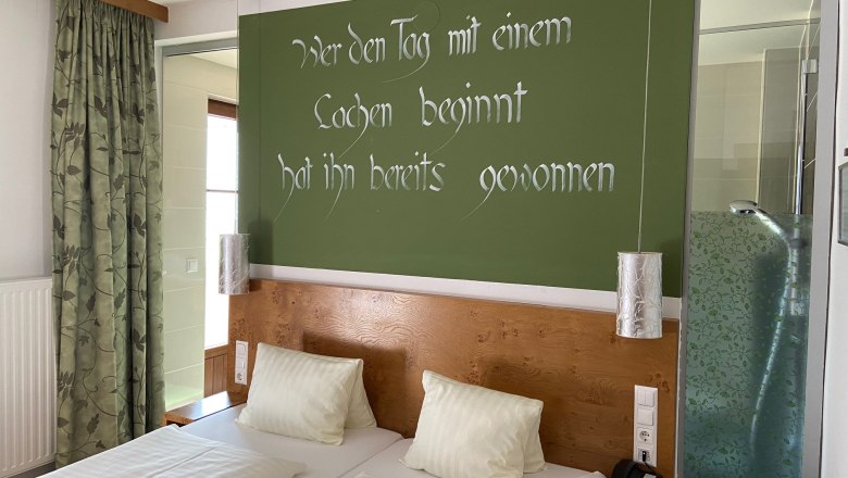 Ein Doppelzimmer mit zwei Betten, gr&uuml;ner Wand mit Spruch, Vorh&auml;ngen und einer Dusche mit Glaswand.