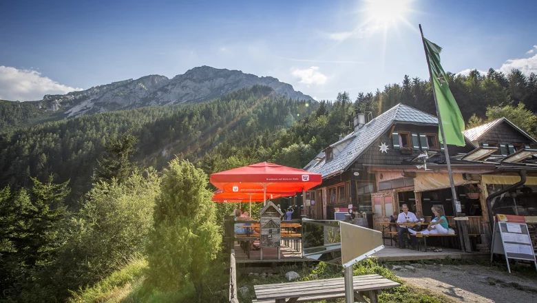 Berggasthof mit Terrasse, roten Sonnenschirmen und Gästen vor bewaldetem Bergpanorama bei Sonnenschein