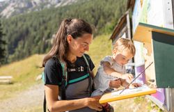 Frau mit Pferdeschwanz und Rucksack hält Kleinkind, das an einer Infotafel im Gebirge zeigt