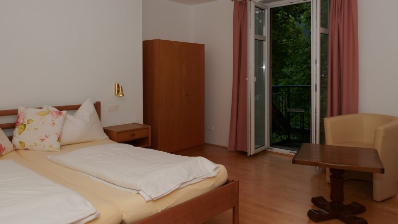 Schlafzimmer mit Doppelbett, Schrank, Tisch und Stuhl, Fenster mit Vorh&auml;ngen.