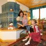 Eine Frau sitzt mit zwei Kindern auf einer Bank vor einem Kachelofen und liest ein Buch. Spielzeug und Dekorationen sind um sie herum verteilt.