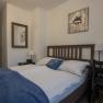 Cozy double room with wooden bed, blue ceiling and decorative cushions. A mirror and a flower picture on the wall, next to it a desk with a plant.